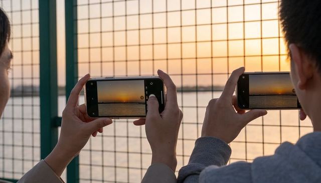 Couple capturing sunset over water holding smartphones framing golden hour seascape