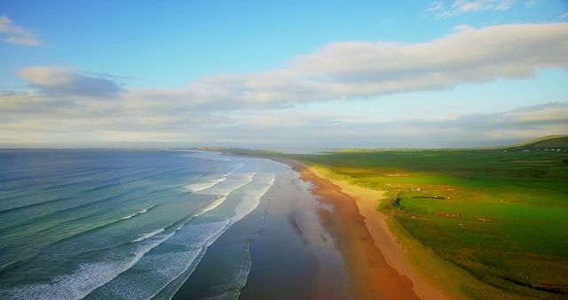 A serene aerial view showcases a sweeping coastline with gentle waves lapping onto the sandy beach, under a vast sky with scattered clouds. The landscape captures the tranquil beauty of a coastal region at what appears to be either dawn or dusk.