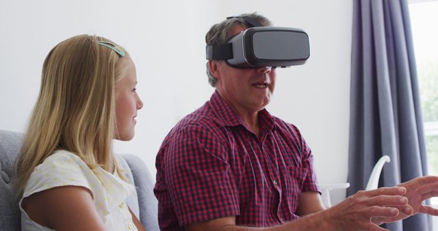 Scene showing an elderly man using a VR headset while being observed by his young granddaughter. Great for illustrating technology bringing generations together, modern family bonding, and the use of innovative devices by older adults.