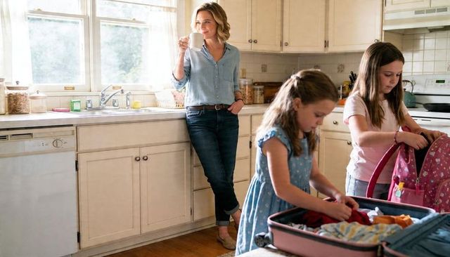 Mother enjoying morning coffee while daughters packing backpacks and suitcase in sunlit kitchen