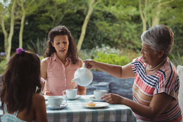 Multigenerational family enjoying tea outdoors in a garden setting. Grandmother pouring tea while mother and child engage in conversation. Ideal for concepts of family bonding, relaxation, and togetherness. Suitable for use in advertisements, family-oriented content, and lifestyle blogs.