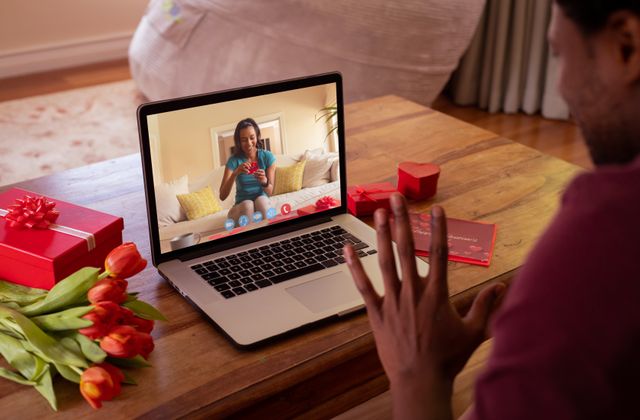 Biracial young woman opening gift while boyfriend waving hand during online dating. unaltered, online dating, video call and distant valentine day celebration.