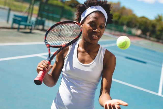 Smiling young african american female player catching ball holding racket at court on sunny day. unaltered, sport, competition and tennis game concept.