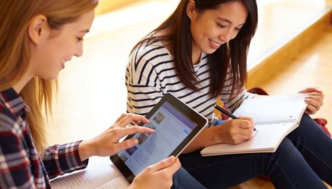 Female students collaborating with tablet and writing notes on campus steps