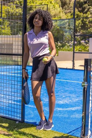 African american woman standing at padel gate holding paddle and ball in lavender polo