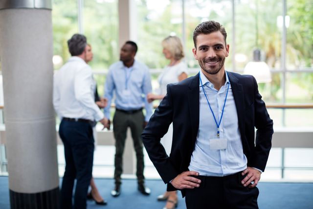 Confident businessman standing with hands on hips, smiling at camera, while colleagues engage in conversation in background. Ideal for use in business-related content, corporate training materials, networking event promotions, and leadership articles.