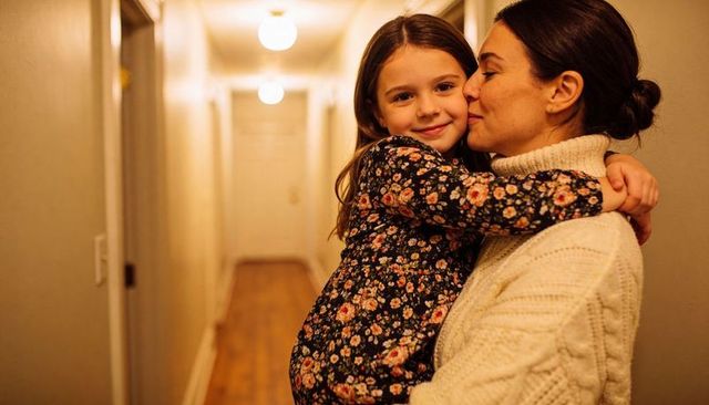 Mother hugging and kissing daughter in cozy hallway, warm home bonding moment with soft lighting