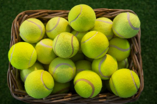 This image shows a wicker basket filled with bright yellow tennis balls placed on a grass field. It is ideal for use in sports-related content, tennis training materials, recreational activity promotions, or articles about outdoor games and fitness.