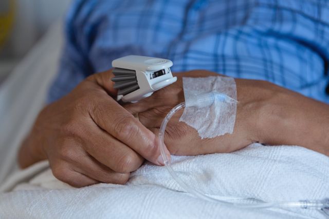 Close-up of a patient's hand with an IV line and pulse oximeter in a hospital setting. Useful for illustrating medical care, patient treatment, healthcare services, and hospital environments. Ideal for health-related articles, medical blogs, and educational materials on patient care and recovery.