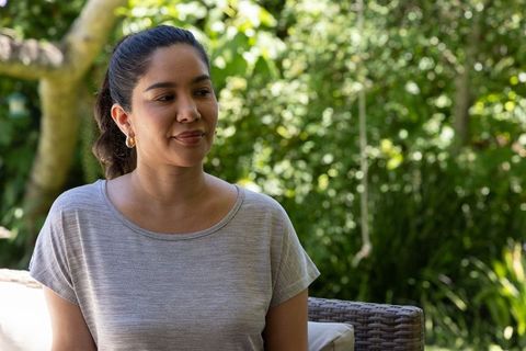Serene woman sitting on wicker chair in sunlit backyard wearing gray knit top and gold hoops