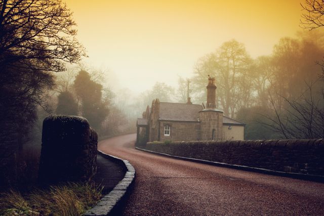 Curving road through tranquil countryside forest enveloped in morning mist with warm sunrise glow. Small house by the roadside creates a picturesque, serene scene. Perfect for use in travel magazines, nature blogs, landscape artwork, or as a calming background in printed materials.