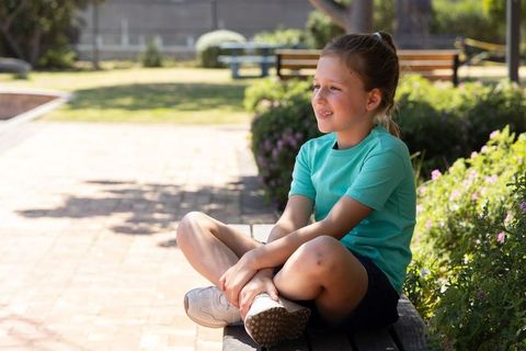 Young girl sitting on bench in sunny park, smiling and relaxing among flowers and greenery