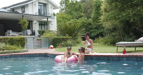 Diverse female friends relaxing on inflatable donut poolside holding drinks and smartphone