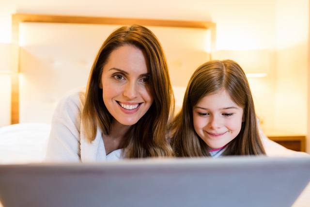 Mother and daughter bonding in cozy bedroom using laptop. Ideal for themes on family time, technology in everyday life, and parent-child relationships. Perfect for articles, blogs, and advertisements focusing on happy family moments, digital learning, and home life.