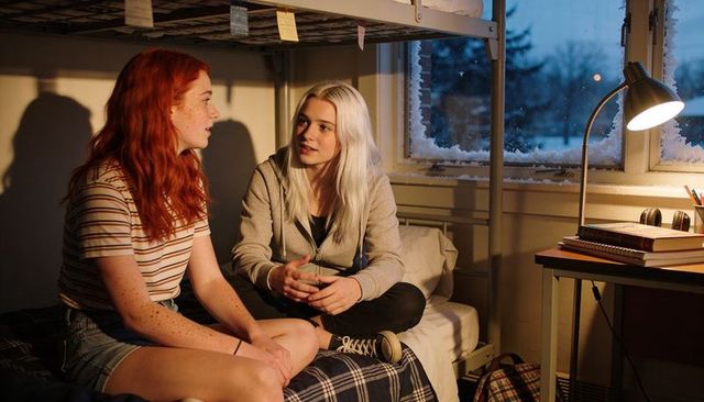 Late-night dorm roommates chatting on bunk bed with frosted window and cozy desk lamp
