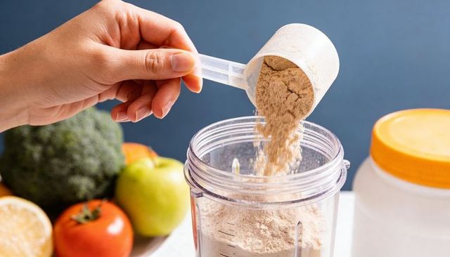 Hand pouring protein powder scoop into blender jar with fresh produce and supplement container