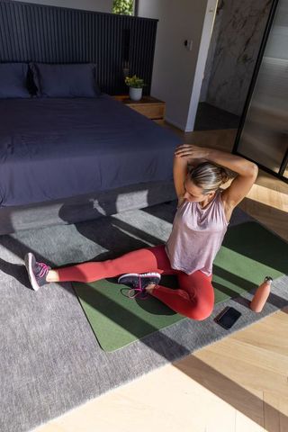 Woman stretching on green yoga mat in bright bedroom morning workout wearing red leggings