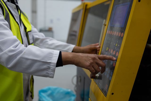 Side view mid section of a Caucasian female worker working in a factory, wearing a lab coat and a high visibility vest, operating a machine on the production line, pushing buttons on a control panel