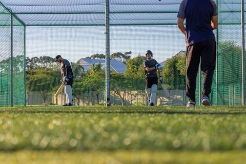Sunlit cricket batting practice in net cage on artificial turf with helmeted players