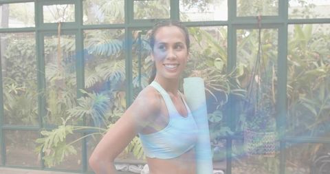 Smiling woman holding rolled yoga mat in bright sunroom with tropical plants