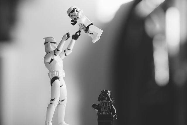 Black and white scene showing a Lego stormtrooper lifting a smaller toy stormtrooper higher, as Darth Vader stands watching. This image captures a playful moment, ideal for themes of family bonding, parenting, or leisure. It can be used for blog posts about parenting in the Star Wars universe, fun themed party invitations, and social media content.