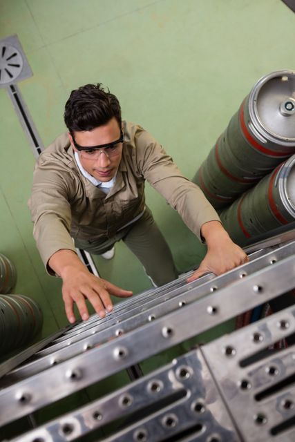 Male worker wearing safety goggles climbing a ladder in a warehouse filled with kegs. Ideal for illustrating industrial work environments, logistics operations, occupational safety, and manual labor. Useful for articles, advertisements, and training materials related to warehouse safety and logistics.