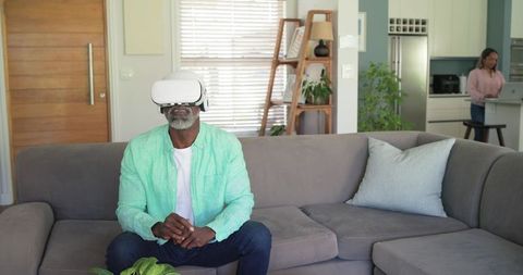 Mature Black man experiencing virtual reality on couch while partner working remotely at home