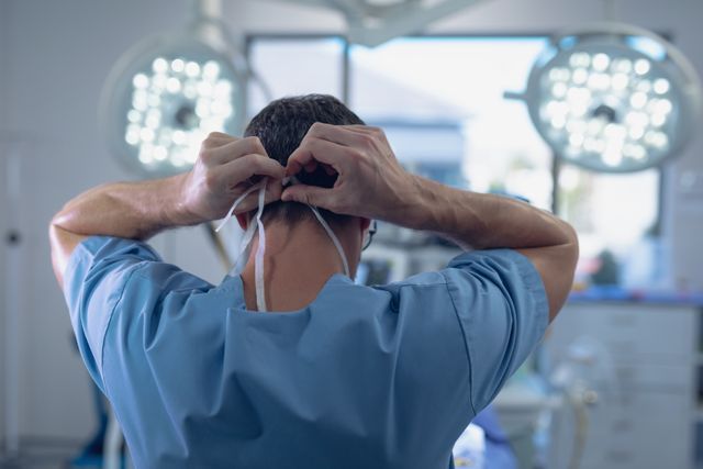 This image captures a rear view of a male surgeon tying a surgical mask in a brightly-lit operation room. It is ideal for use in healthcare-related content, medical websites, hospital brochures, or educational materials emphasizing surgical procedures and medical professionalism.