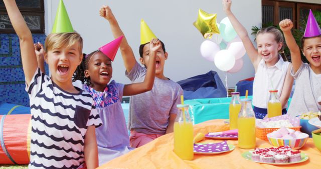 A diverse group of children celebrates a birthday party outdoors, with copy space. They are wearing party hats and cheering with joy around a table with snacks and drinks.