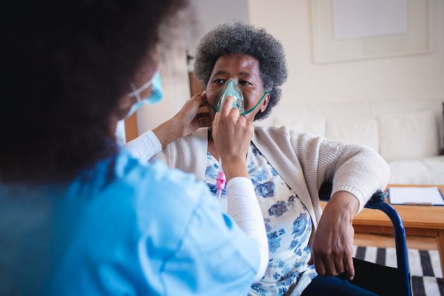 Female doctor putting oxygen mask on african american senior woman sitting at home. senior homecare concept