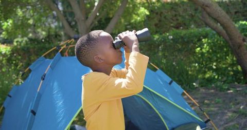 African-american boy exploring sunlit forest using binoculars at blue tent campsite