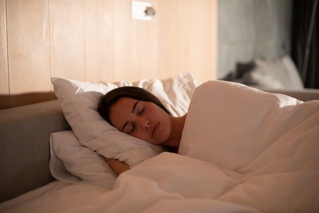 This image depicts a Caucasian brunette woman sleeping peacefully on her side in bed, covered with a white duvet. Ideal for use in articles or advertisements related to sleep health, relaxation, bedroom furniture, bedding products, or wellness. It conveys a sense of tranquility and comfort, making it suitable for promoting sleep aids, mattresses, or nighttime routines.