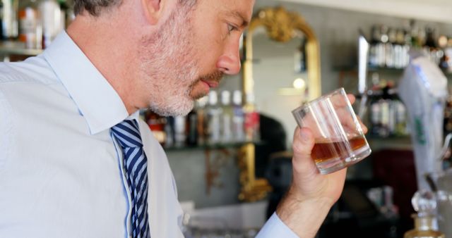 Mature businessman in a bar holding a glass of whiskey, appearing to contemplate. Ideal for use in business-themed publications, content focused on relaxation and winding down after work, or articles related to professional lifestyle and social events.