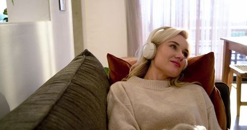 Blonde woman relaxing on sofa with wireless headphones enjoying cozy sunlit living room