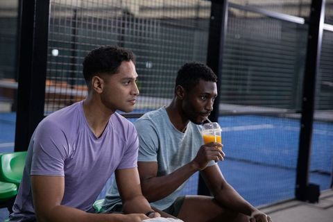 Diverse male teammates sitting courtside on indoor court sharing orange drink and towel