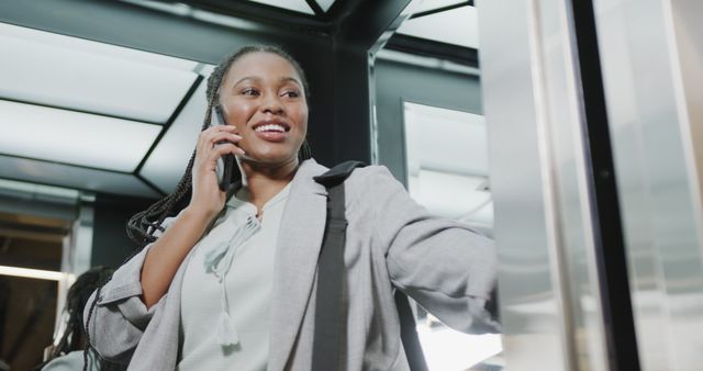 Confident African American businesswoman talking on phone inside modern elevator. Perfect for themes of business communication, corporate life, professional attire, and successful women. Suitable for use in corporate websites, business brochures, and career-related articles.