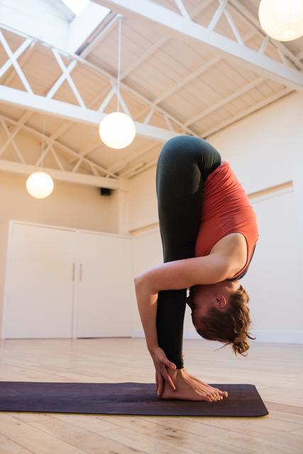 Woman practicing standing forward bend yoga pose in a bright, spacious fitness studio. Ideal for use in wellness blogs, fitness websites, yoga class promotions, and health-related articles.
