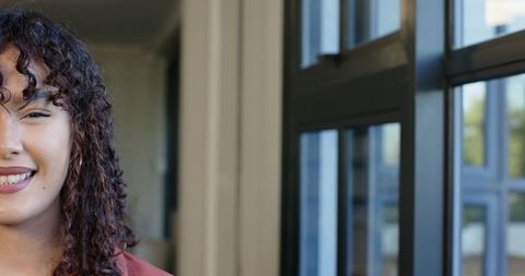 Smiling young woman with curly hair and nose stud by sunlit window with copy space