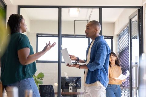 Diverse team discussing project while holding laptop and papers in open-plan glass office
