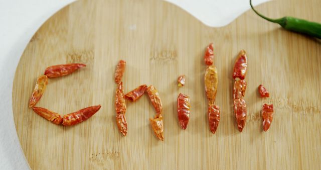 Dried red chili peppers are arranged on a wooden cutting board to spell the word CHILI, with a fresh green chili pepper to the side. The creative display showcases the key ingredient in spicy cuisines, emphasizing its heat and flavor.