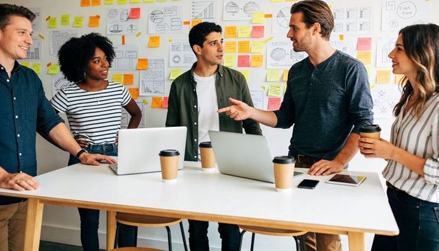Five colleagues brainstorming around standing table with sticky notes, laptops and coffee