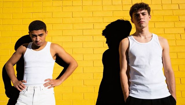 Two young men wearing white tank tops posing on vibrant yellow brick wall, high-contrast