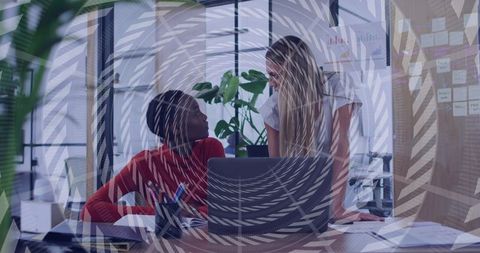 Two women collaborating at glass office desk reviewing laptop and paperwork, teamwork
