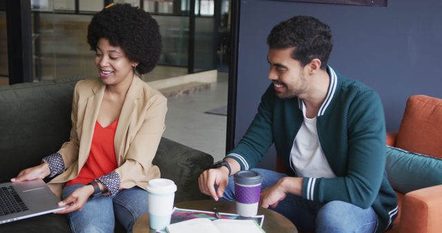 Young African American entrepreneurs are collaborating on a project in a modern office space. They are smiling and using a laptop while having coffee cups on the table, suggesting a casual and productive working environment. This image can be used for business, teamwork, and modern workplace themes in media regarding startups, corporate environments, and entrepreneurial ventures.