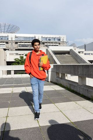 African-american teen walking on urban campus plaza wearing orange hoodie holding notebook
