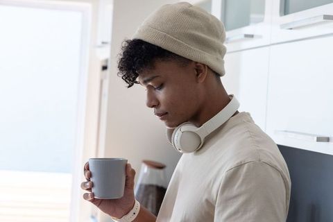 Young man sipping morning coffee in bright kitchen wearing beanie and headphones
