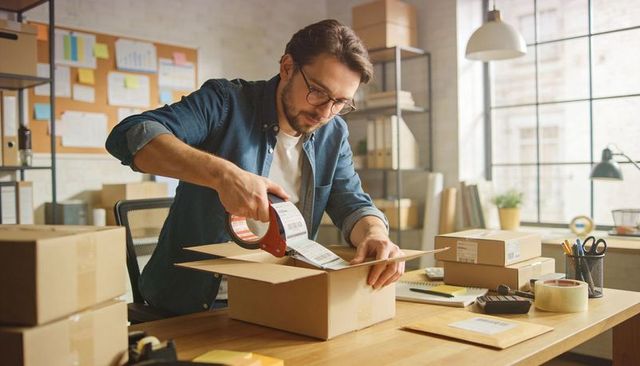 Small Business Owner Packing Orders at Desk Using Tape Dispenser and Shipping Labels for online shop