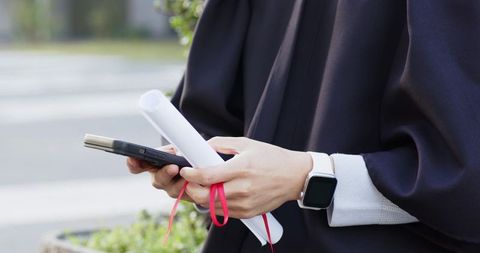 Asian graduate checking smartphone while holding diploma with smartwatch on urban campus