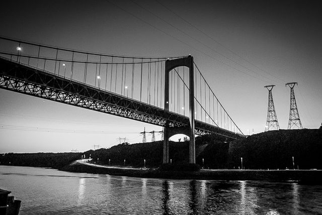 Black and white depiction of a large suspension bridge extending over a calm river at twilight. The lights on the bridge shine brightly, reflecting off the water, and power lines stretch across the sky. Ideal for use in projects related to infrastructure, engineering, transportation, urban planning, and cityscapes.