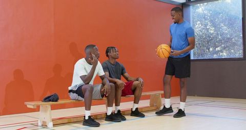 Standing player holding basketball while teammates sitting on bench chatting in indoor court